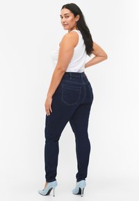 Jeans alta vita di colore blu scuro in denim, con una texture liscia, stile a cinque tasche e cuciture a contrasto. I tacchi completano l'outfit.