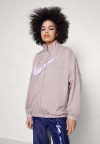 Ljusrosa Nike vindjacka med hög krage, dragkedja och vitt logotyp. Har elastiska ärmslut och en avslappnad passform, tillverkad av lätt material.