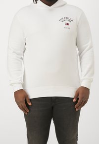 Uomo che indossa un hoodie bianco di Hilfiger New York con logo sul petto e jeans grigio scuro, in piedi con le mani rilassate ai lati.