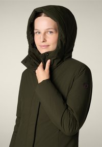 Chaqueta aislante verde oliva con un alto cuello y capucha, que cuenta con un cierre de cremallera y botones a presión, mostrando un diseño elegante y una textura suave.