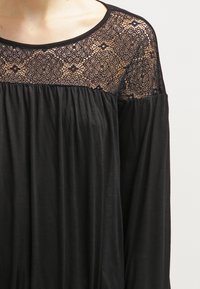 Svart klänning med spetsaxelparti med blomstertryck, långa ärmar och en avslappnad, flödande byst gjord av mjukt tyg.