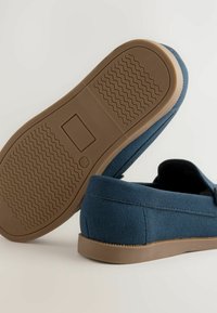 Des chaussures en daim bleu à enfiler avec un bout arrondi et des semelles en caoutchouc marron présentant des motifs de grip texturés. Couture visible le long des bords.
