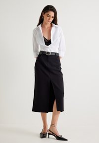 Chemise blanche à manches longues sur un haut en dentelle noir, jupe noire taille haute avec fente avant, et chaussures noires à talons avec des bouts pointus et des embellissements.