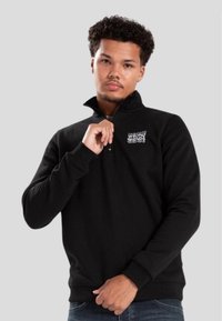 Sudadera negra de forro polar con cuello de media cremallera y texto del logo "WRONG FRIENDS" en el lado izquierdo del pecho. Presenta mangas largas y puños de canalé.
