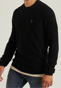 Maglione nero lavorato a maglia con collo rotondo, polsini a costine e un tessuto strutturato. Presenta un piccolo logo sul petto. Indossato sopra una camicia di colore chiaro.