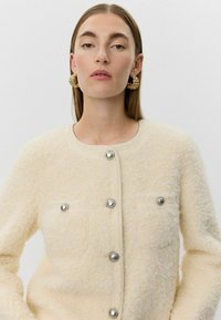 Krémovo sfarbená textúrovaná bunda s kovovými patentmi a dvoma náprsními vreckami. Model má na sebe veľké zlaté kruhové náušnice.