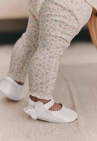 Baltas ādas apavi ar tauriņa akcentu, valkāti ar gaišām ziedu raksta legingām, kas iezīmē pastelīgas krāsas un smalku tekstūru.