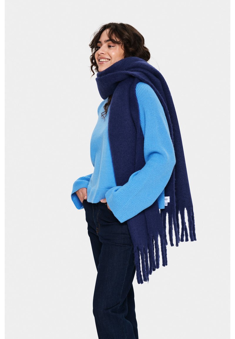 Écharpe bleu marine avec des extrémités frangées, faite d'un matériau doux. Portée par-dessus un pull bleu clair avec une texture côtelée, associée à un jean foncé.