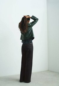 Giacca cropped in similpelle verde con maniche lunghe sopra una maglietta nera, abbinata a jeans scuri a gamba larga a vita alta.