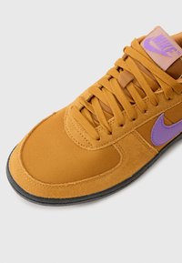 Nike tenisice s gornjim dijelom od sueda i mreže u boji senfa, ljubičastim logom swoosh, ravnim vezicama i crnom gumenom petom.