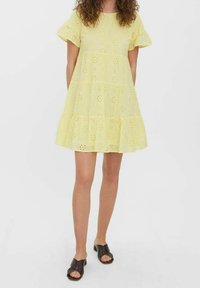 Geel eyelet-jurkje met korte ruches aan de mouwen en een gelaagde rok. Zachte textuur met bloemachtige uitsnijdingspatronen. Gecombineerd met donkere instapschoenen.