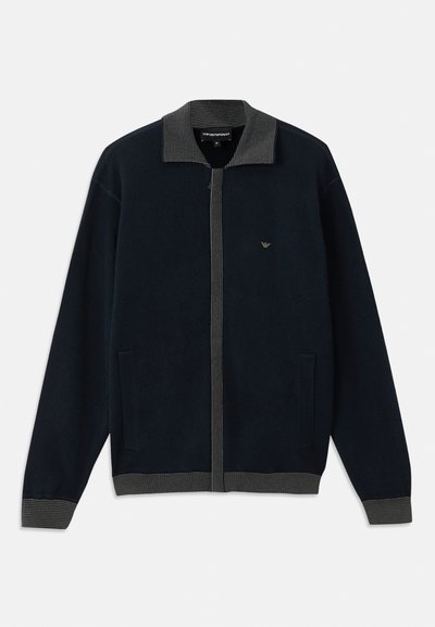 Cardigã azul-marinho escuro com colarinho e punhos canelados. Apresenta um fecho de correr na frente, dois bolsos laterais e um logótipo discreto no peito. Textura suave.