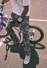 Cargo pants in gray camouflage, paired with gray sneakers. The person is leaning against a purple BMX bike on asphalt.