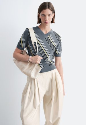 Femme portant un pull gris rayé à col en V et manches courtes, un pantalon plissé crème taille haute, et portant un sac à épaule en cuir crème.