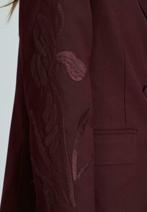 Bordowa marynarka z haftowanym kwiatowym wzorem na rękawie, o gładkiej fakturze i z pojedynczą kieszonką.