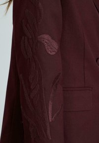 Bordeaux blazer met ingewikkelde kastanjebruine bloemenborduurwerk op de mouwen, met een soepele textuur en een enkele zakdetail.