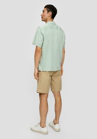 Lichtgroen shirt met korte mouwen en een rechte zoom, gecombineerd met beige shorts. Model draagt witte sneakers, gezien van achteren.