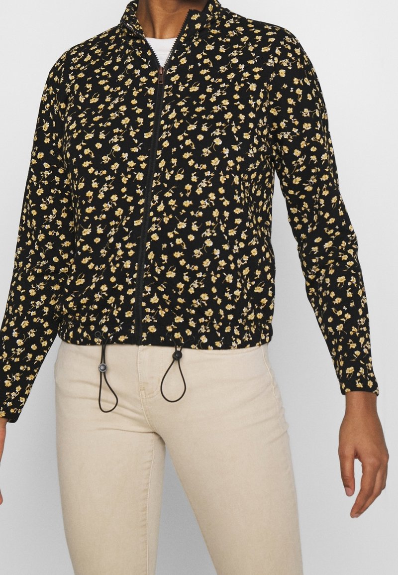 Personne portant une veste noire zippée avec un petit motif floral jaune et blanc, un pantalon beige et une chemise blanche en dessous.