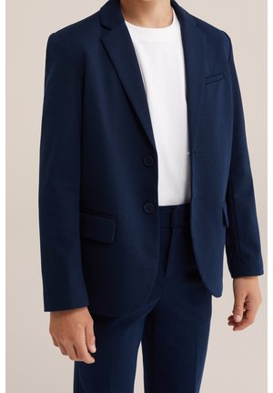 Barn iført en navyblå jakkesætjakke og bukser med en hvid t-shirt under, stående foran en neutral baggrund.