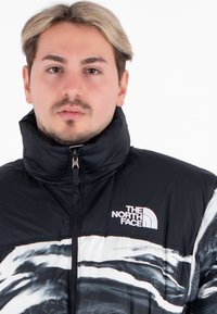 Chaqueta acolchada en blanco y negro con cuello alto, que presenta un cierre con cremallera en la parte delantera y el logo "The North Face" en el pecho, exhibiendo patrones abstractos de olas.