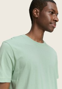 T-shirt verde chiaro a girocollo realizzata in morbido tessuto di cotone; maniche corte e vestibilità morbida, con una texture liscia e dettagli minimi.