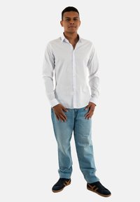 Chemise blanche à manches longues avec un col, associée à un jean bleu clair et des baskets noires avec des accents marron.
