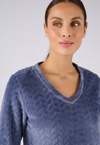 Suéter azul texturizado con cuello en V y un sutil detalle dorado en el escote; hecho de una tela suave con un patrón de trenzas.
