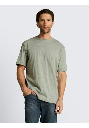 Man met kort krullend haar, gekleed in een lichtgrijs T-shirt met korte mouwen en een blauwe spijkerbroek, staand met één hand in zijn zak.