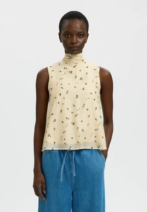 Blusa beige senza maniche con collo alto e piccole stampe floreali nere, abbinata a pantaloni di jeans blu larghi con vita regolabile con coulisse.