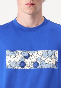 Modré bavlněné tričko s obdélníkovou grafikou, která obsahuje bílý a světle modrý text v kresleném stylu a ikony úsměvů. Kolo krku.
