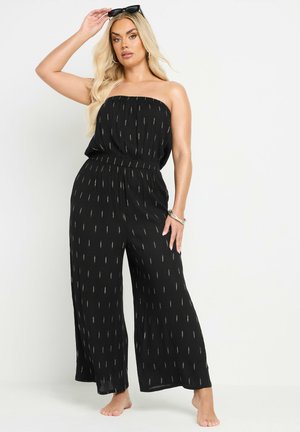Mustat olkaimettomat jumpsuitit, joissa on kultaisia pystysuoria kuvioita, elastinen vyötärö, leveälahkeinen muotoilu ja kevyet kankaat. Mallilla ei ole kenkiä.
