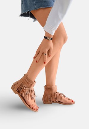 Jambes portant des sandales à franges couleur beige, une jupe en jean, une manche blanche, une montre noire, une bague argentée, et du vernis à ongles rouge sur les doigts et les orteils.