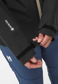 Black waterproof jacket features adjustable Velcro cuffs. The material is smooth with a GORE-TEX label prominently displayed.