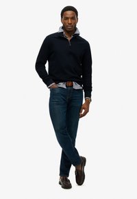 Mørkeblå zip-neck trøje over en lyseblå skjorte med krave, parret med mørkeblå jeans og brune loafers. Inkluderer et brunt bælte og et ur.