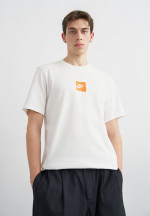 Νεαρός άνδρας που φοράει λευκό μπλουζάκι Nike με πορτοκαλί λογότυπο και μαύρο παντελόνι, στέκεται μπροστά σε απλό φωτεινό φόντο.