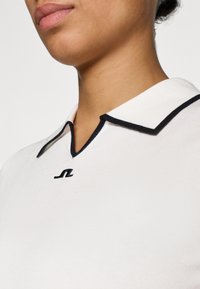 Λευκό πον κι με γιακά, με μαύρη ρίγα και κεντημένο λογότυπο, στενή γραμμή, λεία υφή, αναδεικνύει την περιοχή του �λαιμού και των ώμων.