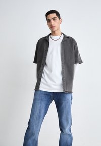 Camisa de botones gris de manga corta sobre una camiseta blanca, combinada con jeans azules. Textura suave, diseño casual, sin patrones ni acentos visibles.