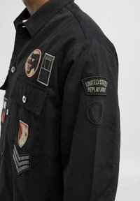 Sort skjorte med flere broderede patches, der har forskellige designs og teksturer. Indeholder en frontlomme og knaplukninger.