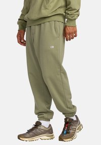 Pantalones de chándal verde oliva con puños y cinturilla elásticos, que cuentan con un pequeño parche de logo en la parte frontal. Combinados con unas botas de senderismo de color marrón claro.