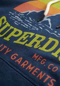 Sudadera azul marino con un gráfico en amarillo y naranja, que presenta montañas y olas. El texto dice "SUPERDRY MFG. CO." en letras gruesas y en contraste.