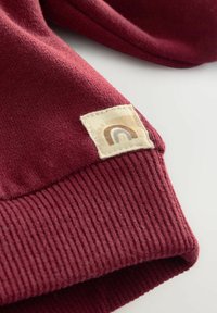 Bordeauxfarbener Sweatshirtbund mit strukturiertem Rippenabschluss, ausgestattet mit einem quadratischen cremefarbenen Stoffpatch mit einem braunen Regenbogenmuster. Weiches Material.