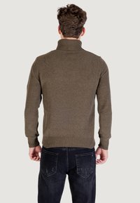 Uomo con capelli ricci visto da dietro, indossa un maglione a collo alto marrone e jeans neri su uno sfondo neutro.