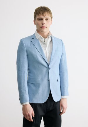 Jeune homme portant un blazer bleu clair sur une chemise blanche à rayures et un pantalon noir, debout devant un fond blanc uni.