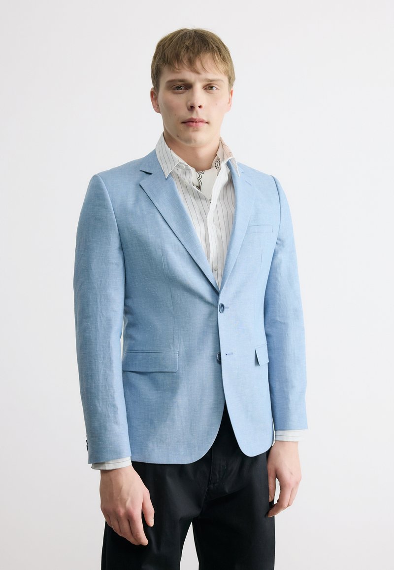 Νέος άνδρας που φοράει ένα ανοιχτό μπλε σακάκι πάνω από ένα λευκό πουκάμισο με ρίγες και μαύρο παντελόνι, στέκεται μπροστά σε ένα απλό λευκό φόντο.