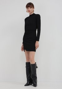 Eine Frau trägt ein enganlegendes schwarzes langärmeliges Mini-Kleid mit einem kleinen Schlitz vorne und schwarzen knielangen Lederstiefeln. Sie steht vor einer weißen Wand.