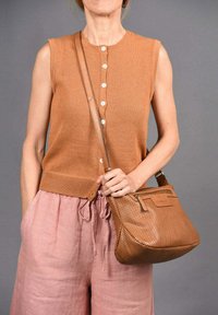 Bruine gebreide mouwloze top met knopen, gecombineerd met losse roze linnen broek, terwijl ze een gestructureerde bruine schoudertas met perforaties vasthoudt.