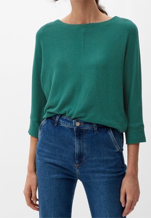 Kobieta w zielonym topie z długim rękawem, wpuszczonym w wysokie niebieskie dżinsy, na tle gładkiej białej ściany.