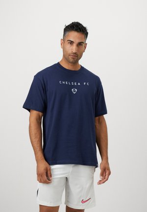 Mand iført navy Chelsea FC t-shirt og hvide shorts med pink Nike-logo, stående foran en ensfarvet lys baggrund.
