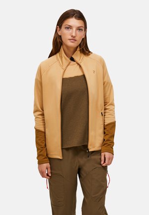 Lichtbeige jas met een hoge kraag, Contrasterende bruine mouwen en ritszakken. Draagt over een bruine top, gecombineerd met groen-bruine broek.