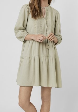 Woman wearing a loose, light beige knee-length dress with long sleeves, front panel embroidery, and tassel ties at the neckline.